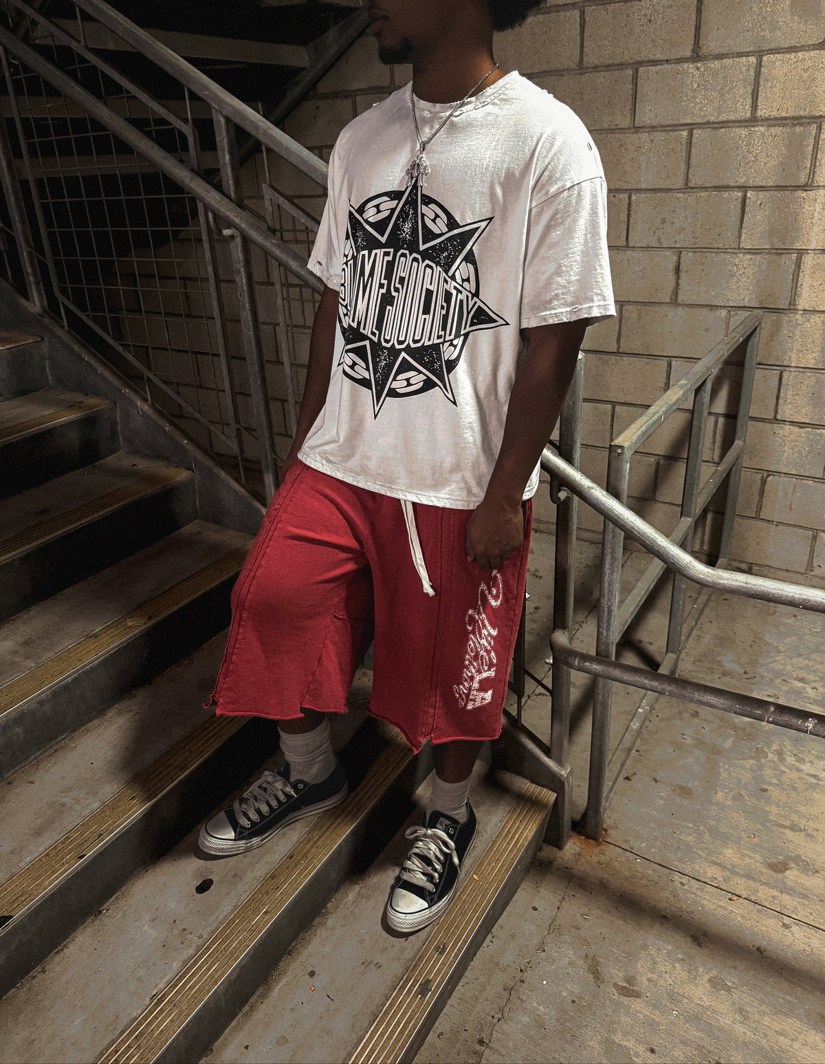 A man wearing a white graphic t-shirt and red shorts stands on concrete stairs in an industrial setting with brick walls and metal railings.